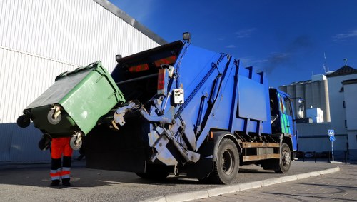 A blue waste collection truck with a hydraulic lifting mechanism, positioned on an asphalt surface next to a white industrial building under a partly cloudy sky. The truck is actively engaged in collecting a large green wheeled bin, which is being lifted and emptied into the truck's hopper. There is a person wearing red work trousers and an orange hi-vis jacket standing nearby, observing the process. The background includes modern industrial structures, and the scene conveys a professional rubbish collection operation consistent with waste management services offered by companies like Commercial Waste West Byfleet, serving the local area near Surrey postcode KT14.