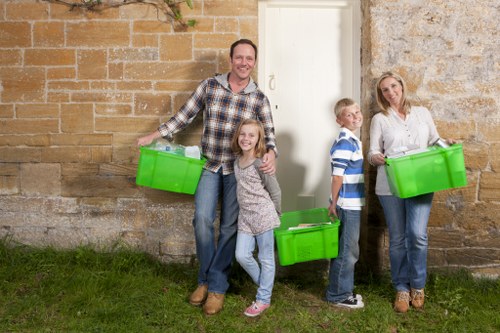 A family of four, including a man, woman, and two children, standing outdoors against a weathered stone wall near a white door, each holding a bright green plastic rubbish bin. The man, on the left, has short brown hair and is wearing a checkered shirt with jeans, smiling at the camera while holding a bin. The young girl next to him, with long light brown hair, is wearing a grey patterned top and jeans, also smiling and holding a bin. To the right, the boy with short blonde hair is dressed in a blue and white striped shirt and jeans, looking slightly to the side, holding his bin ready for rubbish collection. The woman, on the far right, has long blonde hair and is wearing a white blouse and jeans, smiling gently while holding her rubbish bin. The scene takes place on a grassy area beside a brick wall, likely near a residential property in West Byfleet, with natural daylight illuminating the scene. This image relates to a rubbish collection or waste management service by Commercial Waste West Byfleet, emphasizing household rubbish disposal.