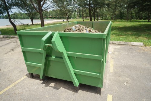 A large green industrial skip with corrugated metal sides and a smooth finish, filled with mixed waste materials including paper, plastic, and organic rubbish. The skip is situated on a paved parking area, with yellow painted lines marking spaces, adjacent to a grassy area with a row of trees. In the background, a lake or river with a low stone or concrete embankment is visible, along with a clear sky and distant greenery. The skip is positioned close to the curb, indicating a waste collection or disposal point, and appears to be part of a commercial waste removal service in West Byfleet, Surrey. The image reflects a typical rubbish removal setup, suitable for commercial or industrial waste clearance, managed by companies like Commercial Waste West Byfleet.